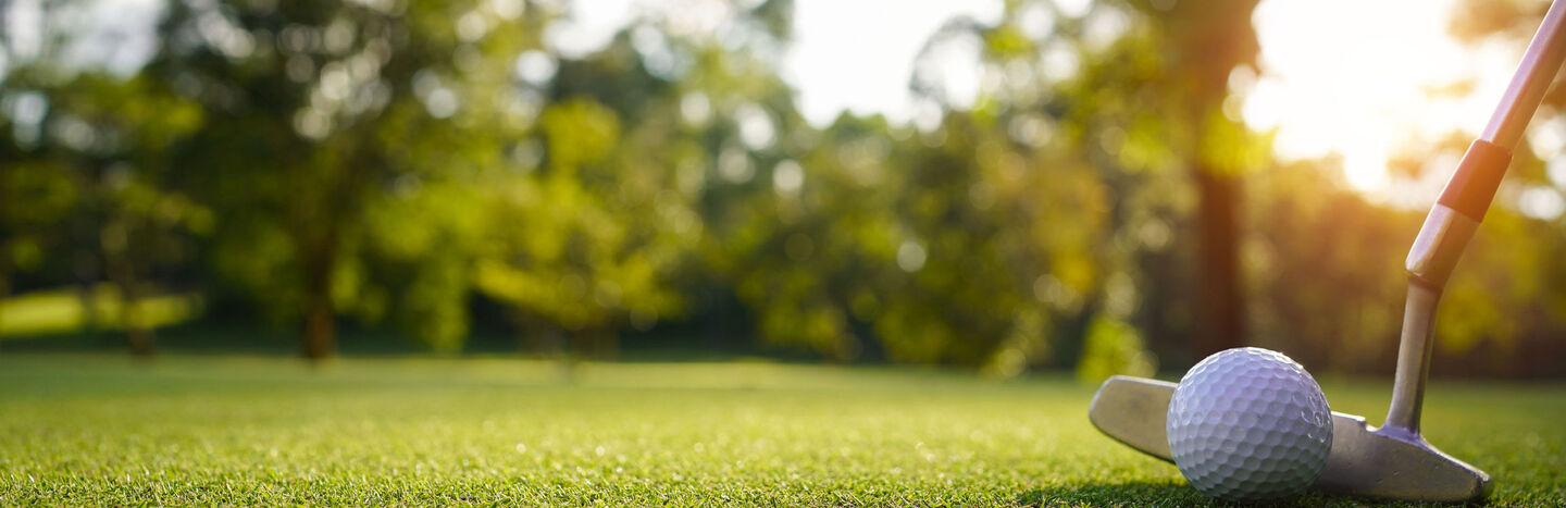 Nahaufnahme eines Golfballs und eines Putters am Rande eines Lochs auf einem Grün mit Bäumen im Hintergrund.