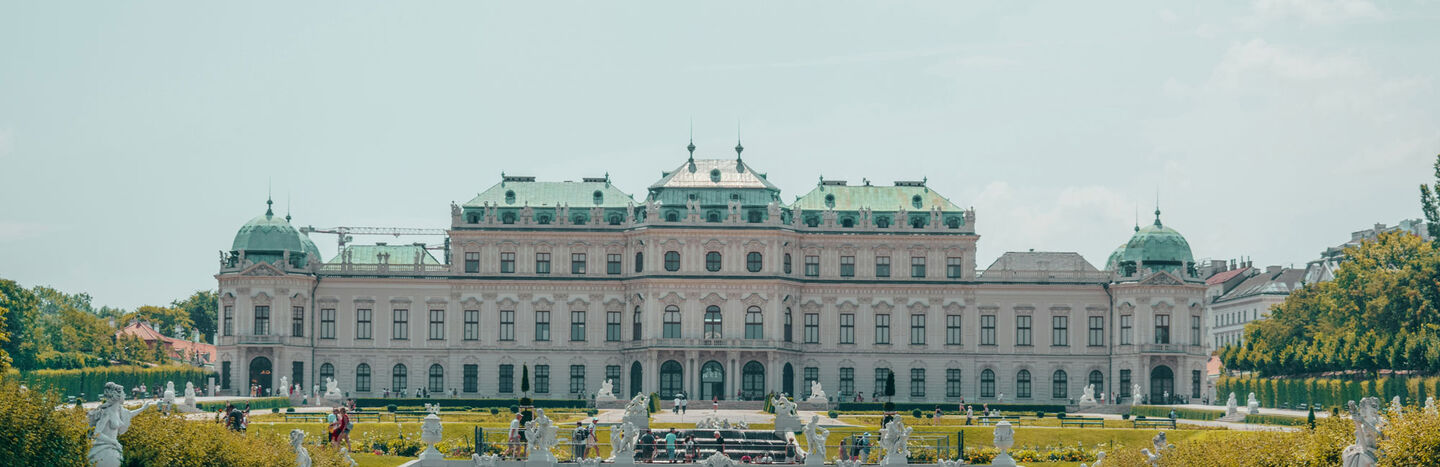 Ein prächtiger Palast mit grünen Kuppeln, formalen Gärten und Statuen unter einem blassblauen Himmel.