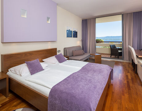 Modernes Hotelzimmer mit Doppelbett, violetten Akzenten, Sofa, Schreibtisch und Balkon mit Blick auf das Wasser und die Hügel.