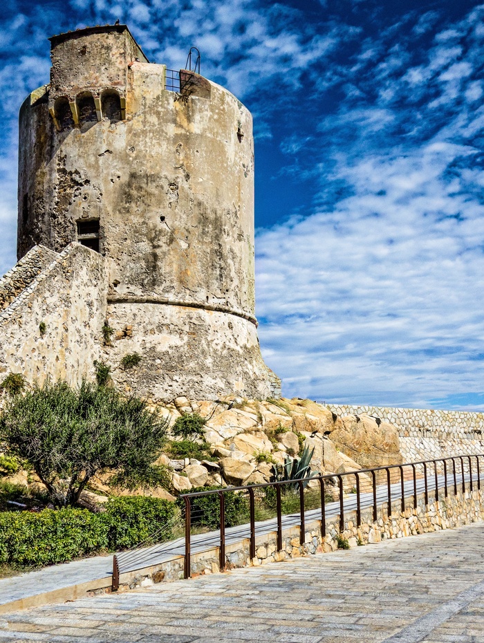 Alter steinerner Wachturm auf einem felsigen Hügel unter einem teilweise bewölkten blauen Himmel, mit einem Gehweg und Geländer davor.