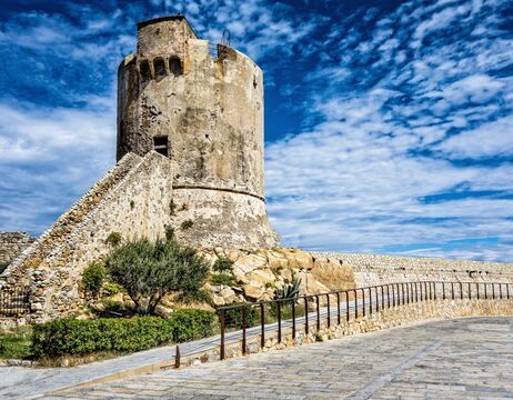 Alter steinerner Wachturm auf einem felsigen Hügel unter einem teilweise bewölkten blauen Himmel, mit einem Gehweg und Geländer davor.