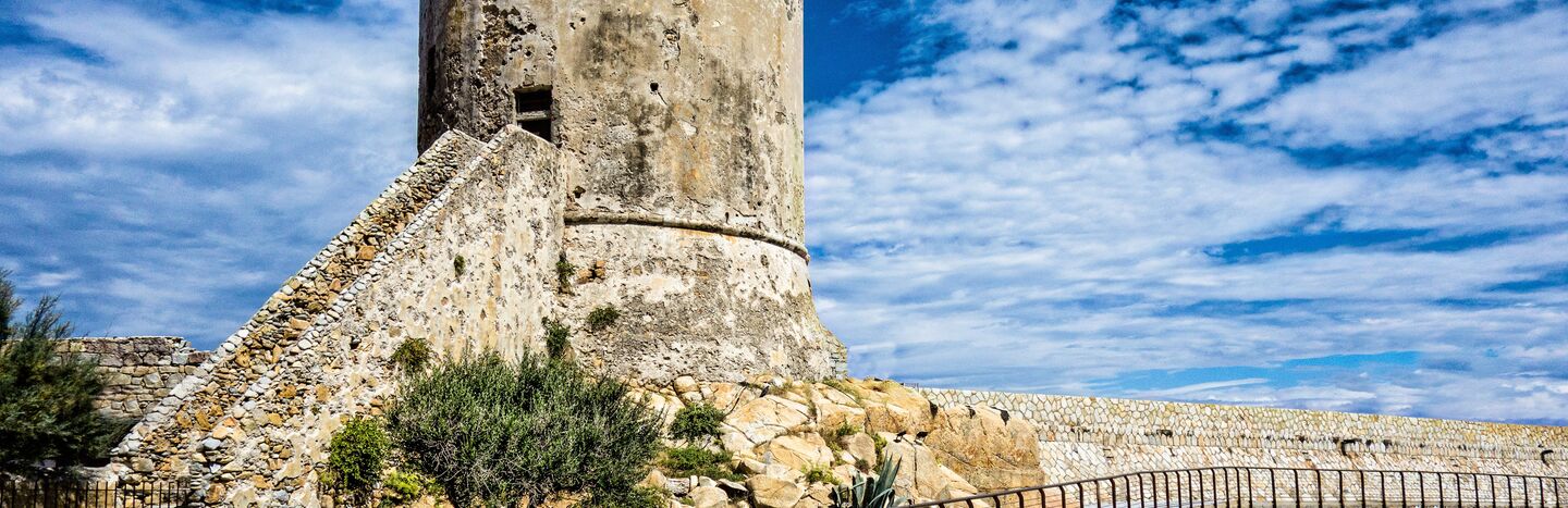 Alter steinerner Wachturm auf einem felsigen Hügel unter einem teilweise bewölkten blauen Himmel, mit einem Gehweg und Geländer davor.
