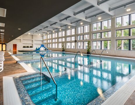 Hallenbad mit großen Fenstern, blauem Wasser, Beckenstufen und einer kleinen Rutsche im Hintergrund.