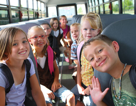 Eine Gruppe lächelnder Kinder mit Rucksäcken sitzt zusammen in einem Schulbus und schaut in die Kamera.
