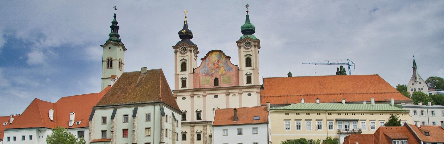 Eine Stadt am Flussufer mit bunten Gebäuden und einer großen Kirche mit Zwillingstürmen im Hintergrund.