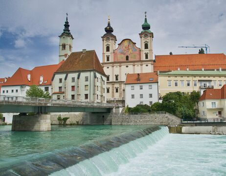 Eine Stadt am Flussufer mit bunten Gebäuden und einer großen Kirche mit Zwillingstürmen im Hintergrund.