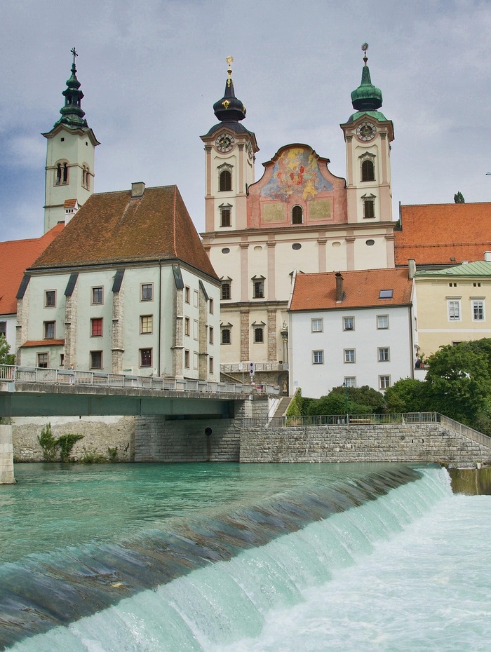 Eine Stadt am Flussufer mit bunten Gebäuden und einer großen Kirche mit Zwillingstürmen im Hintergrund.