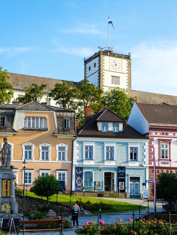 Bunte Häuser und ein Uhrenturm unter blauem Himmel, mit einer Statue und Grün im Vordergrund.
