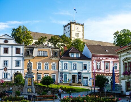 Bunte Häuser und ein Uhrenturm unter blauem Himmel, mit einer Statue und Grün im Vordergrund.