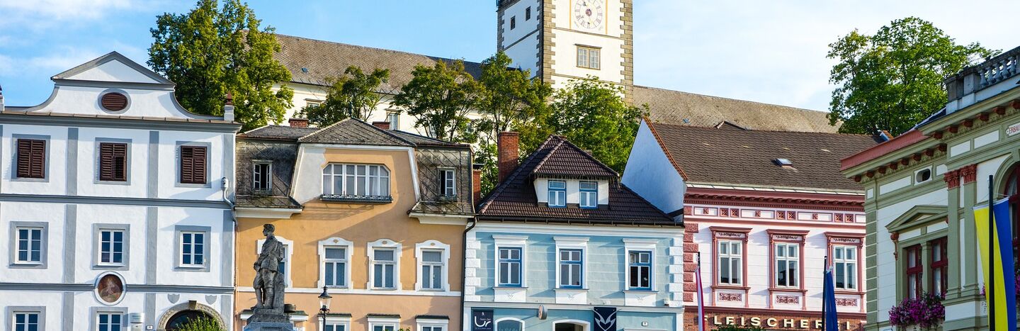 Bunte Häuser und ein Uhrenturm unter blauem Himmel, mit einer Statue und Grün im Vordergrund.