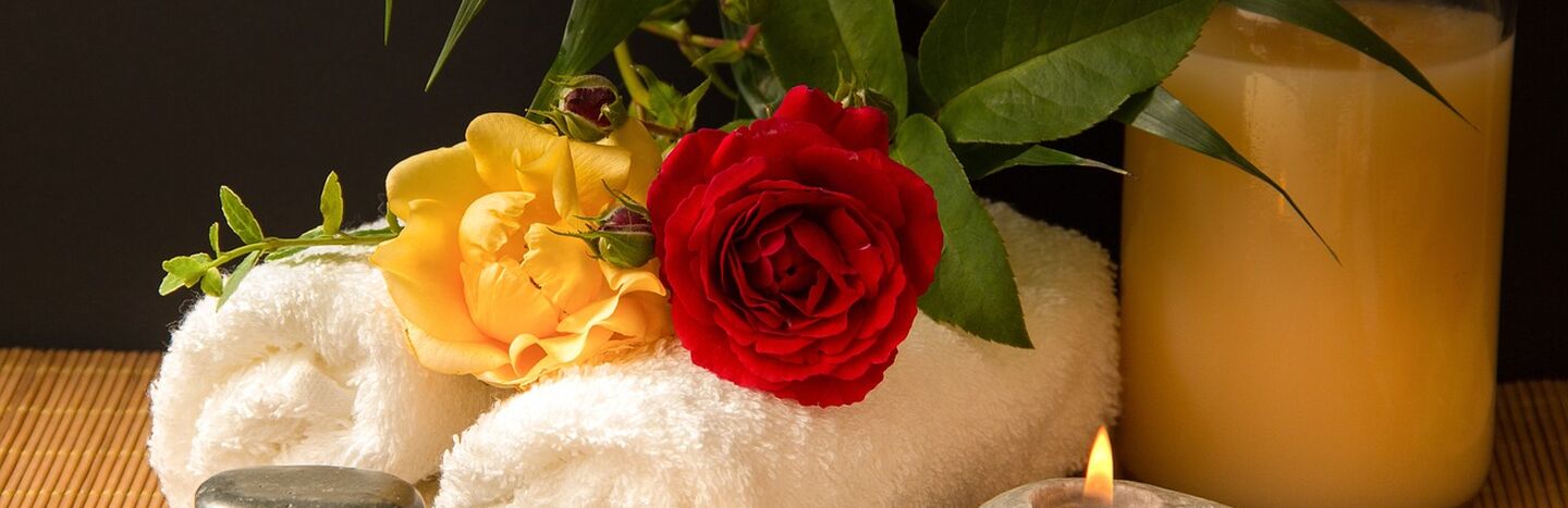 Aufgerolltes weißes Handtuch mit einer roten und gelben Rose, einer Kerze und grünen Blättern, die ein entspannendes Spa-Ambiente schaffen.