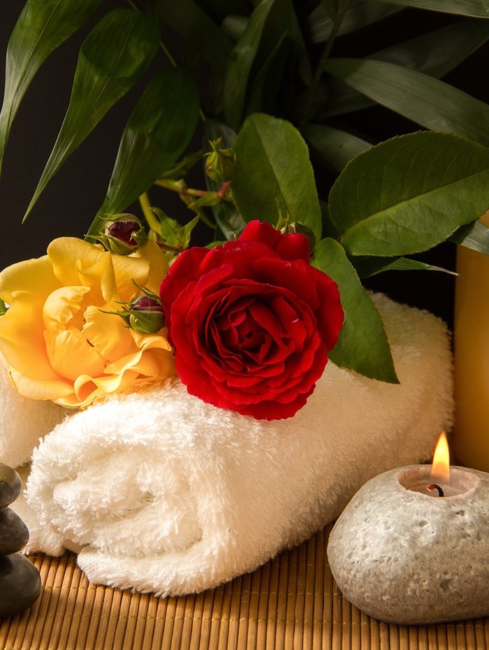 Aufgerolltes weißes Handtuch mit einer roten und gelben Rose, einer Kerze und grünen Blättern, die ein entspannendes Spa-Ambiente schaffen.