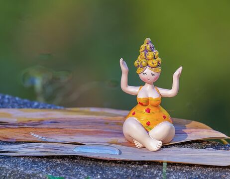 Eine kleine Figur in einem gelben Kleid sitzt im Schneidersitz mit erhobenen Armen auf einer hölzernen Unterlage im Freien.