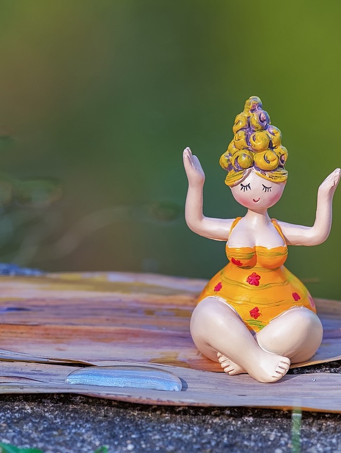 Eine kleine Figur in einem gelben Kleid sitzt im Schneidersitz mit erhobenen Armen auf einer hölzernen Unterlage im Freien.