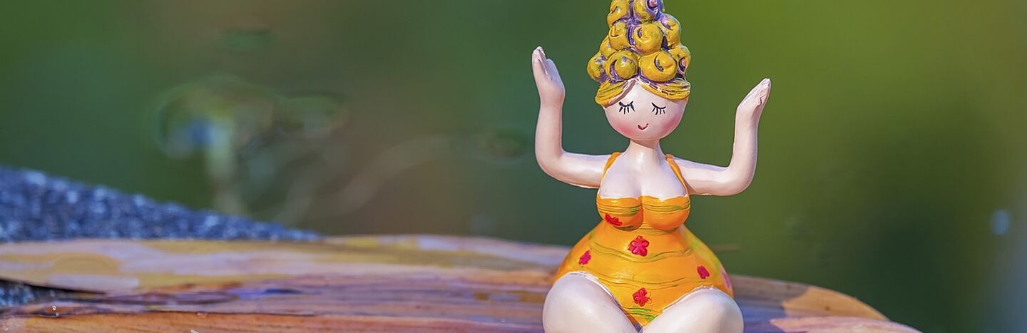 Eine kleine Figur in einem gelben Kleid sitzt im Schneidersitz mit erhobenen Armen auf einer hölzernen Unterlage im Freien.